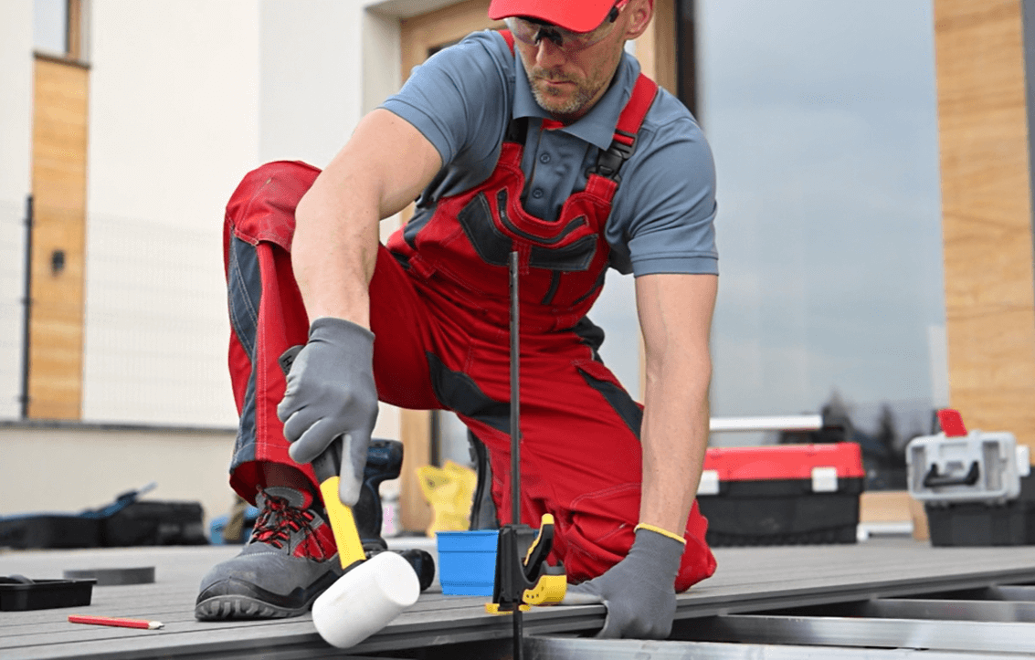 Handwerker in roter Arbeitskleidung montiert Bambus Terrassendielen auf einer Aluminium Unterkonstruktion mit einem weißen Gummihammer; im Hintergrund Werkzeugkoffer und modernes Gebäude.