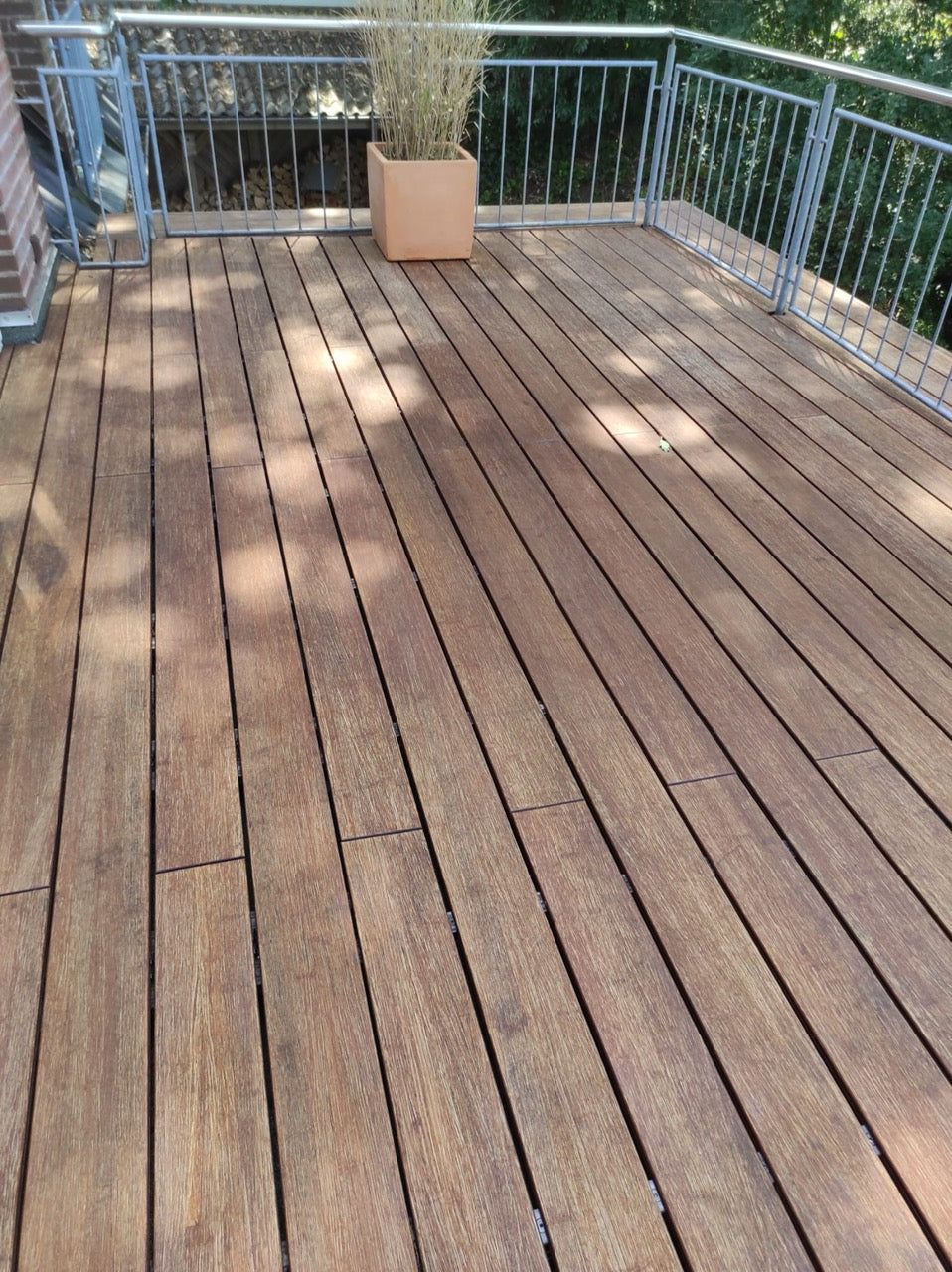 Vorher-Nachher-Vergleich einer Holzterrasse mit Edelstahlgeländer. Links sind verwitterte, graue Terrassendielen vor der Reinigung oder Pflege zu sehen, rechts dieselbe Terrasse nach der Aufbereitung mit deutlich aufgefrischter, warmbrauner Holzoberfläche. Die Terrasse befindet sich an einem Wohnhaus und ist von Bäumen umgeben.