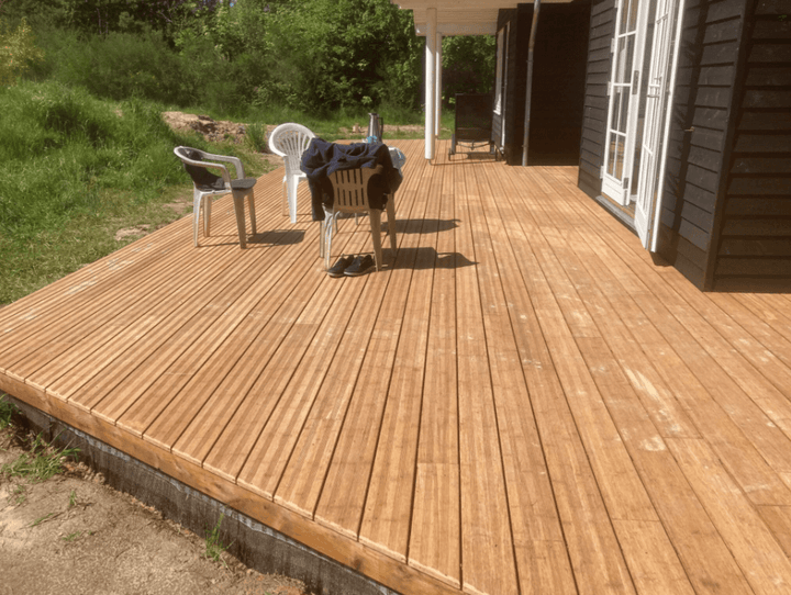 Großzügige Bambusterrasse aus glatten Terrassendielen an einem dunkel verkleideten Haus mit überdachtem Sitzbereich. Auf der Terrasse stehen mehrere Kunststoffstühle, im Hintergrund befinden sich Grünflächen und Bäume
