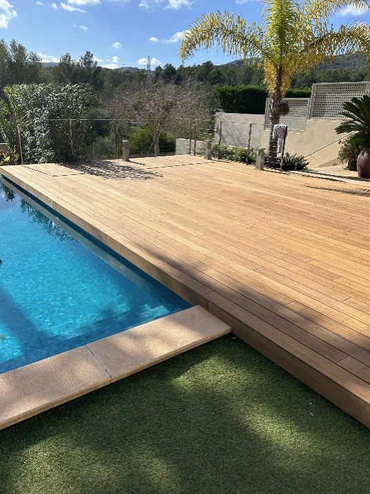 Eine weitläufige Terrasse aus hellen Sand Holzbambusdielen erstreckt sich entlang eines Swimmingpools mit klarem, blauem Wasser. Die Terrasse wird von Palmen, Sträuchern und Bäumen umgeben und schafft eine moderne, mediterrane Atmosphäre im Garten.