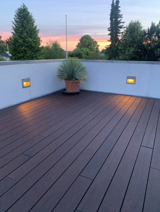 Eine moderne Dachterrasse mit dunkelbraunen Terrassendielen aus Bambus erstreckt sich bis zu einer weißen Brüstungsmauer. Zwei in die Wand integrierte Leuchten spenden warmes Licht. In der Ecke steht eine große Topfpflanze, während im Hintergrund Bäume und ein farbenfroher Sonnenuntergang zu sehen sind.