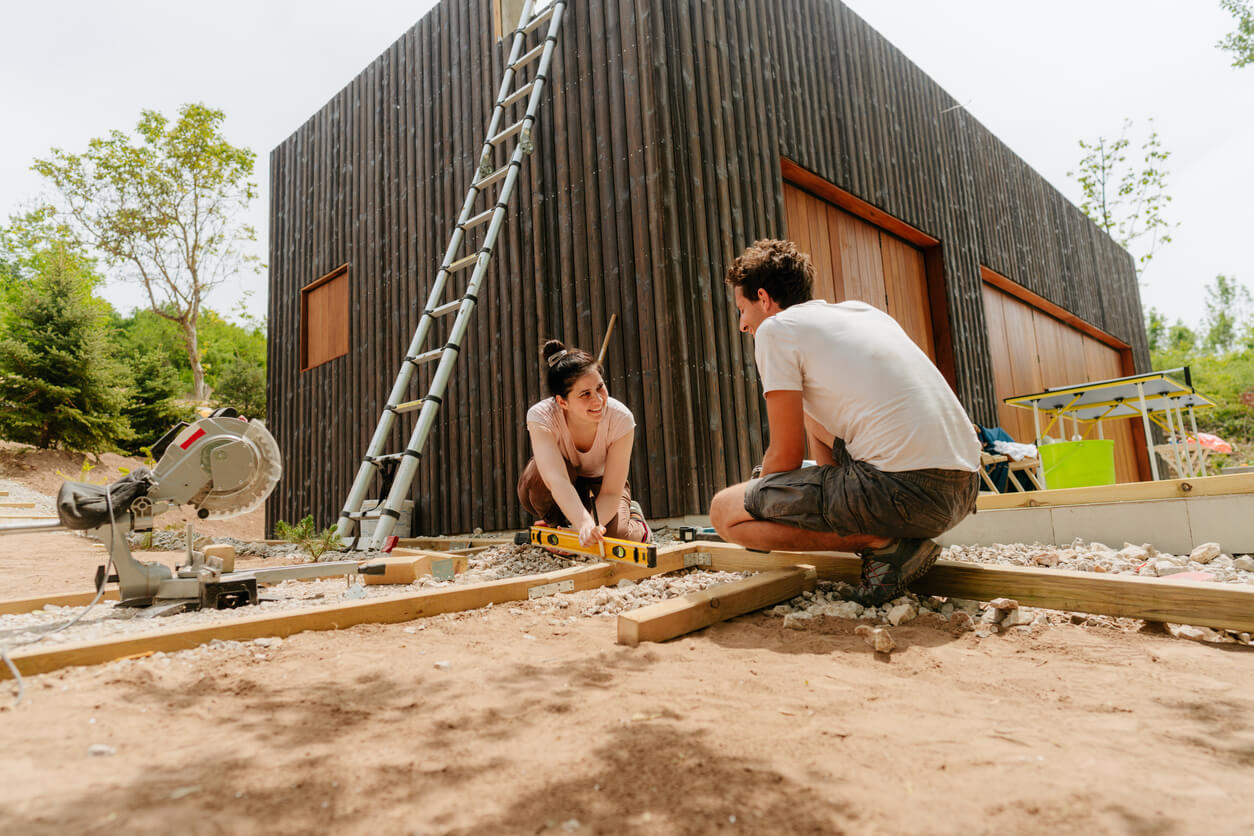 Ein Paar bereitet gemeinsam den Unterbau für ein  aMbooo Terrassen Komplettpaket vor. Auf einem verdichteten Splittbett werden Holzträger mit einer Wasserwaage ausgerichtet – die Basis für die spätere Verlegung von langlebigen Bambus-Terrassendielen.