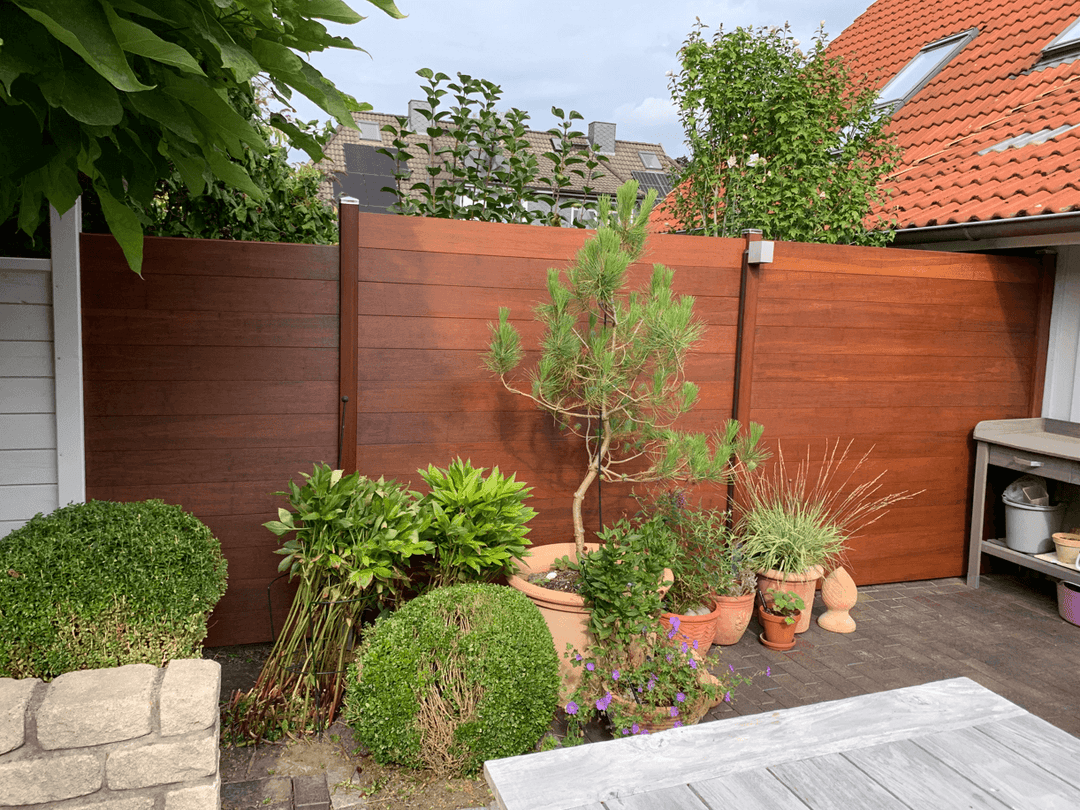 Moderner aMbooo Bambus Sichtschutzzaun in warmer Optik mit dunklen Pfosten, der eine Terrasse begrenzt und als Hintergrund für diverse Gartenpflanzen und einen Pflanztisch dient.