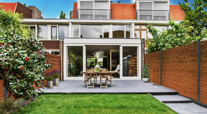 Blick in einen modernen Hinterhof: Eine geflieste Terrasse mit Holztisch und -stühlen vor einer großen Glasfront. Rechts und im Hintergrund begrenzt ein hoher Bambus Sichtschutzzaun mit horizontalen, braunen Latten den Garten. Davor liegt eine gepflegte Rasenfläche, umgeben von grünen Büschen und Pflanzen