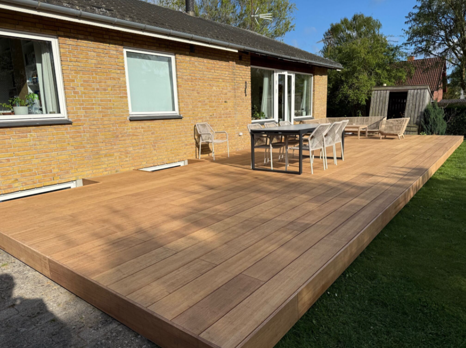 Großzügige Terrasse aus Closed Decking aMbooo Bambus Terrassendielen in natürlicher Holzoptik, ausgestattet mit modernem Essbereich und Lounge-Möbeln, vor einem Einfamilienhaus mit gelber Klinkerfassade – pflegeleicht und wetterbeständig.
