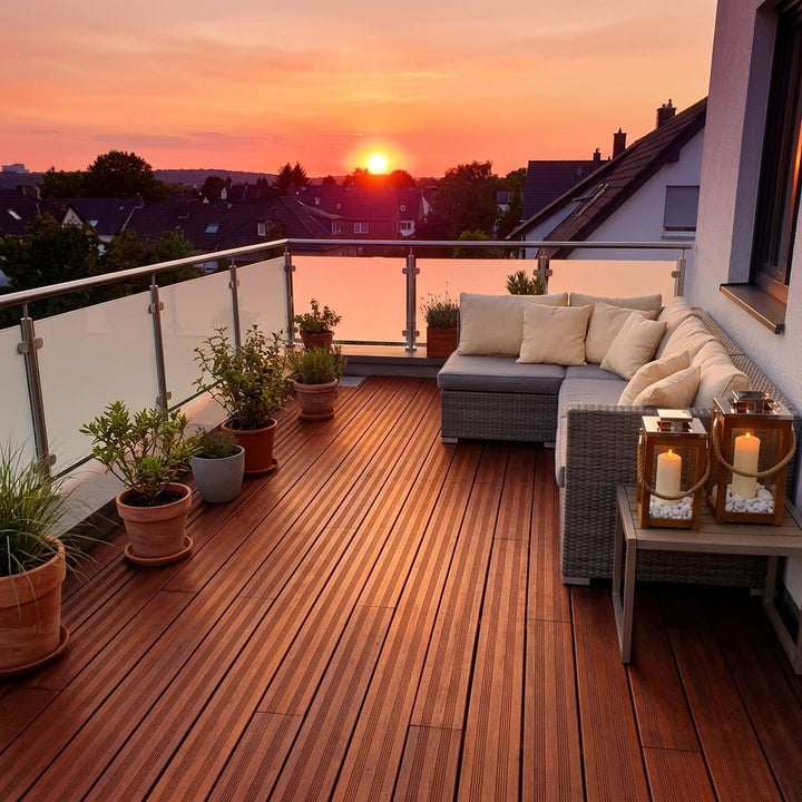 Moderner Balkon mit Holzdielen, Glasgeländer, Sitzmöbeln und Kübelpflanzen im warmen Licht des Sonnenuntergangs.