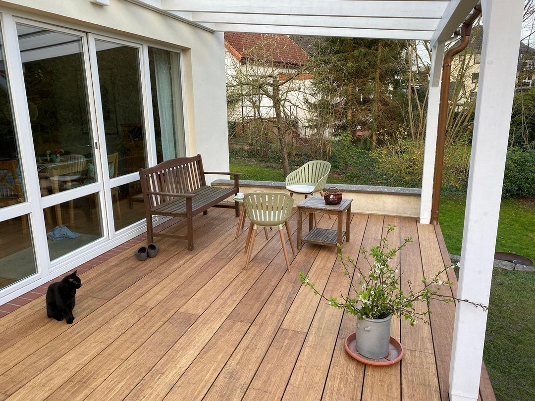 Moderne Terrasse mit hochwertigen glatten XL Bambus Terrassendielen, in der Farbe Sand  unter einer weißen Pergola. Gemütlicher Sitzbereich mit Gartenblick direkt am Wohnhaus mit großer Fensterfront.