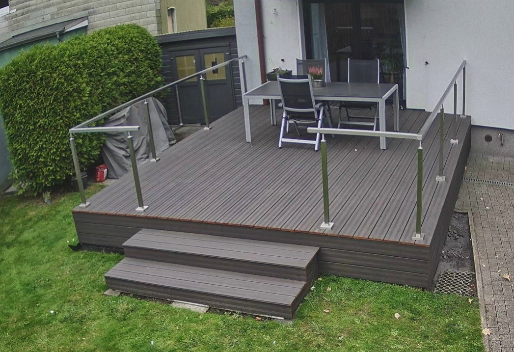 Moderne Terrasse in Grau, vor dem Oelen, mit breiter Treppe, Edelstahlgeländer und Zugang zum Haus. Die Terrasse liegt ebenerdig und grenzt an einen gepflegten Garten.“