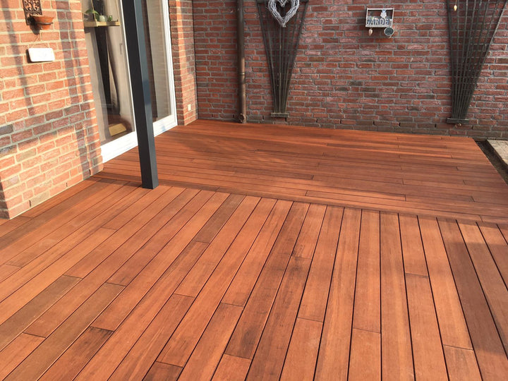 Großzügige Bambusterrasse aus rötlich-braunen Terrassendielen an einer Backsteinfassade, teilweise überdacht von einer modernen Pergola mit dunklen Pfosten. Die Dielen sind längs verlegt und wirken gepflegt, während Sonnenlicht und Schattenmuster über die Fläche fallen. Im Hintergrund befinden sich weitere Backsteinwände, Pflanzgefäße und ein Gartenbereich.