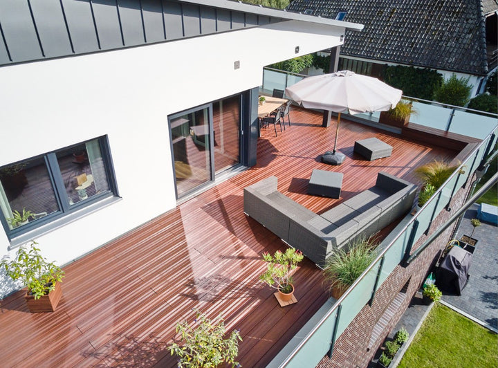 Moderne Dachterrasse mit edlen Holz-Terrassendielen mit glatter Oberfläche, gemütlicher Sitzlandschaft und Sonnenschirm. Das Glasgeländer bietet einen freien Blick auf die umliegenden Gärten und Häuser.