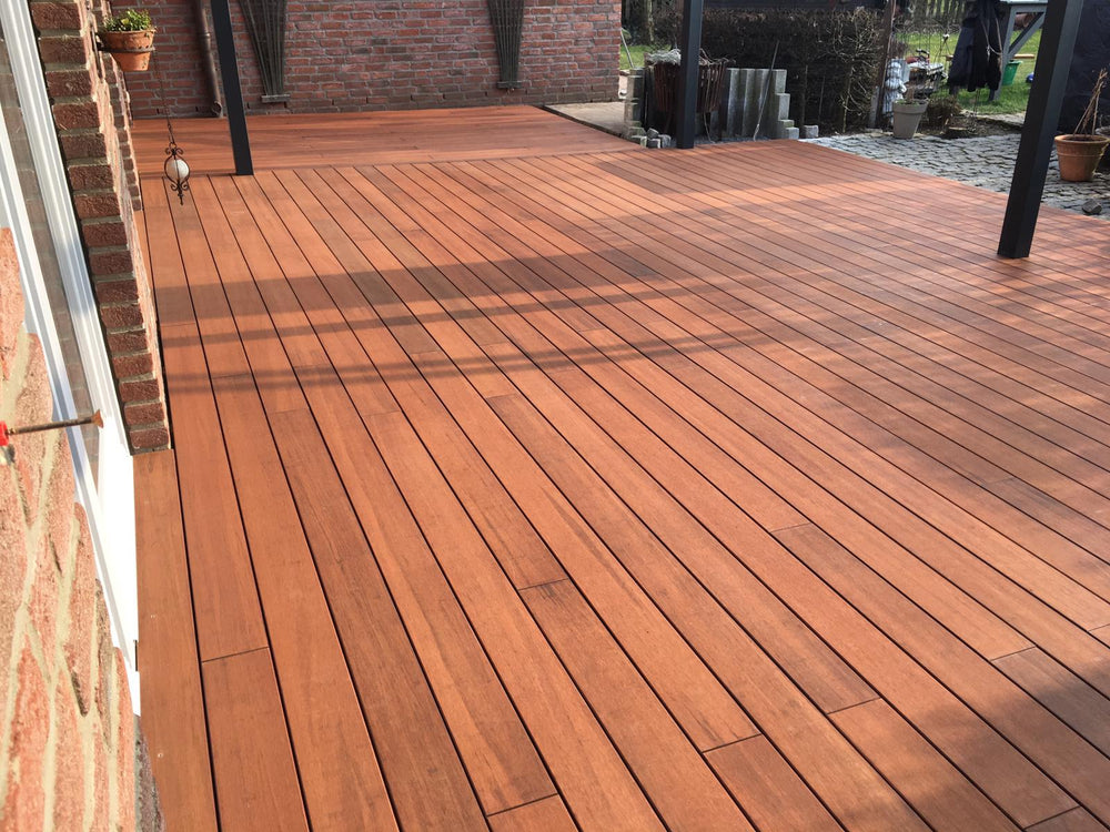 Großzügige Bambusholzterrasse aus rötlich-braunen Terrassendielen an einer Backsteinfassade, teilweise überdacht von einer modernen Pergola mit dunklen Pfosten. Die Dielen sind längs verlegt und wirken gepflegt, während Sonnenlicht und Schattenmuster über die Fläche fallen. Im Hintergrund befinden sich weitere Backsteinwände, Pflanzgefäße und ein Gartenbereich.