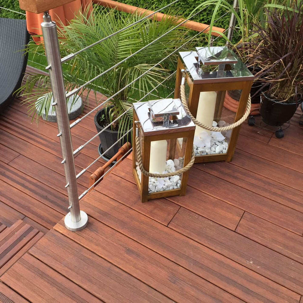 Detailaufnahme einer AMBOOOTerrasse aus rötlich-braunen Holzdielen mit moderner Edelstahlseil-Brüstung. Zwei dekorative Holzlaternen mit Kerzen und weißen Ziersteinen stehen zwischen verschiedenen Kübelpflanzen und verleihen dem Außenbereich eine stilvolle, mediterrane Atmosphäre.