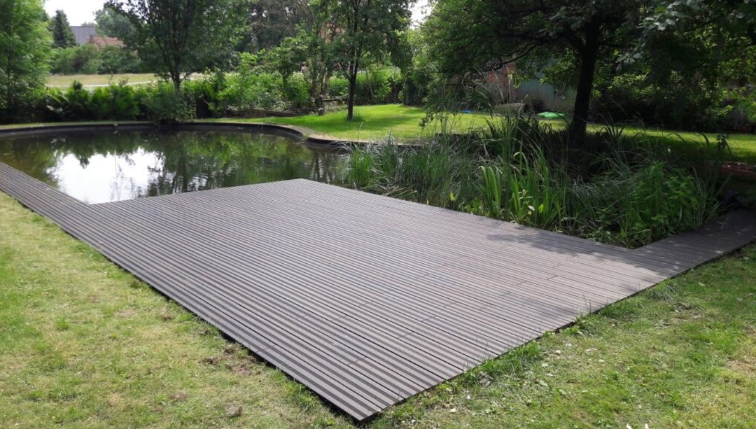 weitläufige Terrasse mit schwarzen, fein geriffelten Terrassendielen erstreckt sich entlang eines naturnah gestalteten Gartenteichs. Das ruhige Wasser spiegelt die umliegenden Bäume und Sträucher wider. Rund um den Teich sorgen Rasenflächen, Schilfpflanzen und dichtes Grün für eine harmonische Gartenatmosphäre. Die Terrasse bietet einen direkten Zugang zum Wasser und fügt sich nahtlos in die natürliche Umgebung ein.