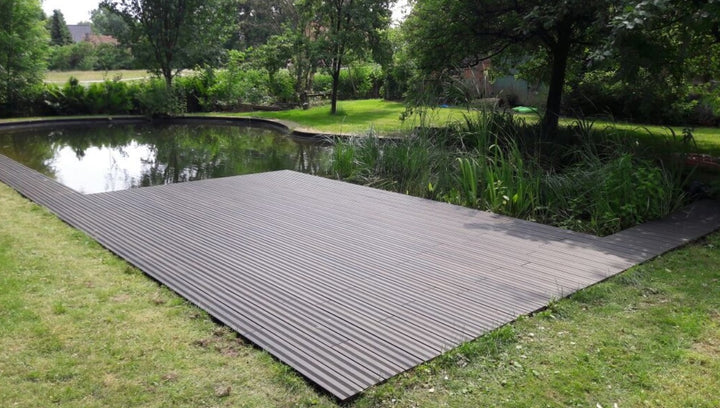 weitläufige Terrasse mit schwarzen, fein geriffelten Terrassendielen erstreckt sich entlang eines naturnah gestalteten Gartenteichs. Das ruhige Wasser spiegelt die umliegenden Bäume und Sträucher wider. Rund um den Teich sorgen Rasenflächen, Schilfpflanzen und dichtes Grün für eine harmonische Gartenatmosphäre. Die Terrasse bietet einen direkten Zugang zum Wasser und fügt sich nahtlos in die natürliche Umgebung ein.